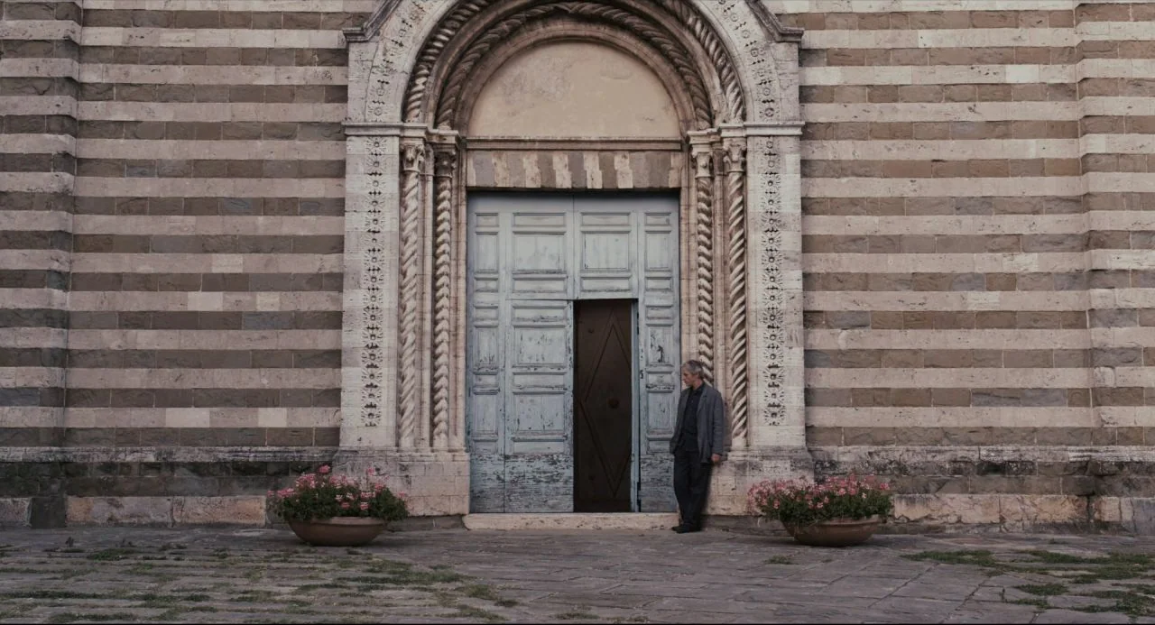 Certified Copy (2010) movie still: wide — A stone building facade features alternating horizontal bands of pale and dark stone blocks. A man stands…
