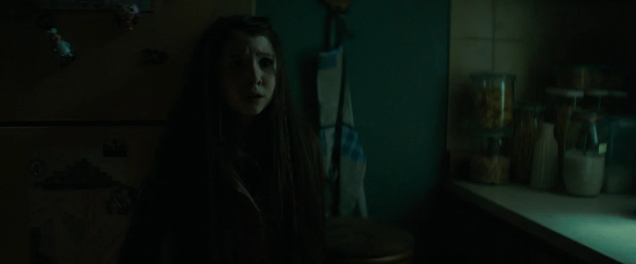 Don’t Breathe 2 (2021) movie still: medium — A dimly lit room with dark teal walls and a cluttered counter with glass jars. A young girl with long…