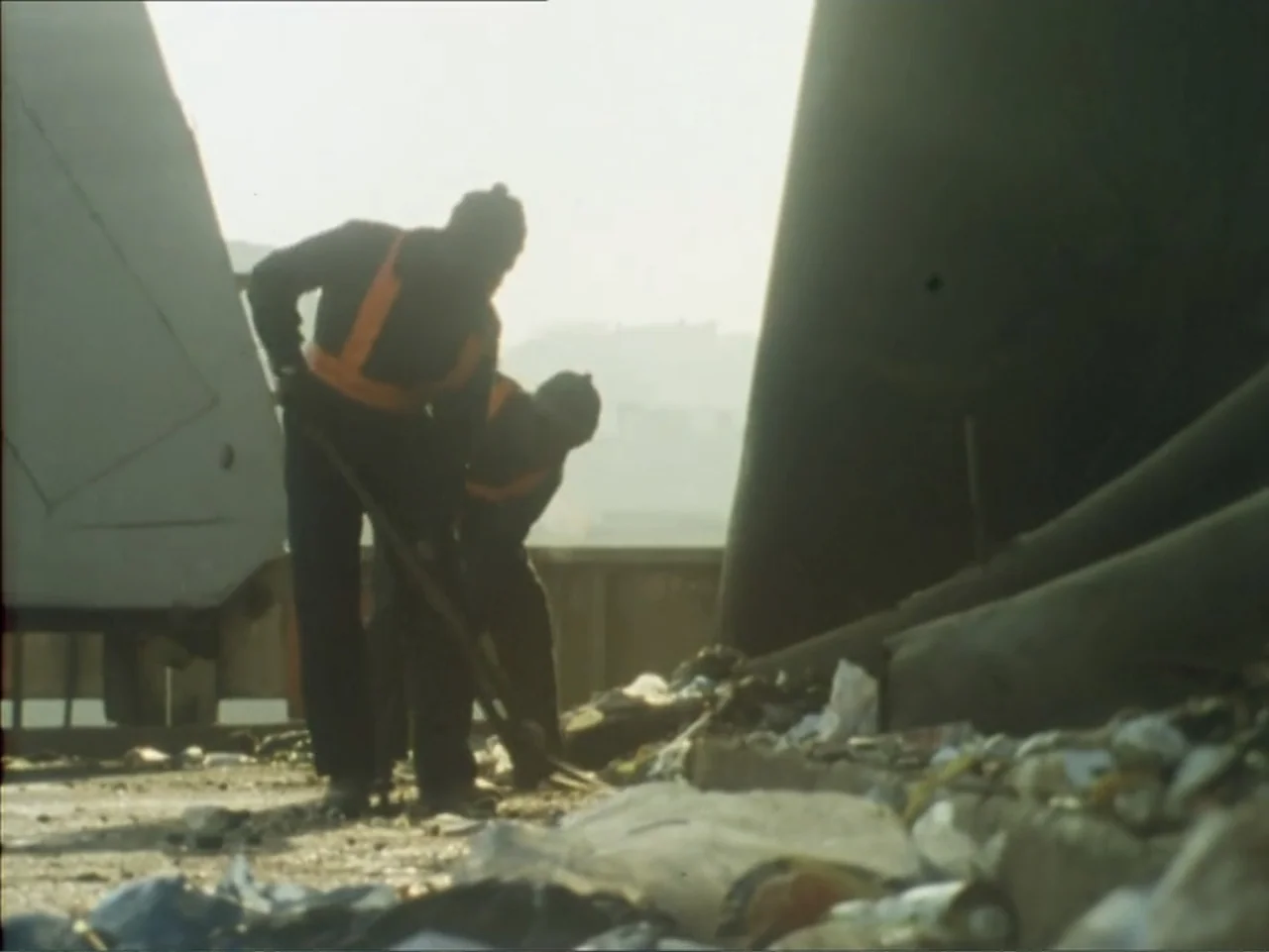 A Dessert for Constance (1981) movie still: medium — An outdoor area is filled with piles of discarded refuse and debris. Two workers in protective…