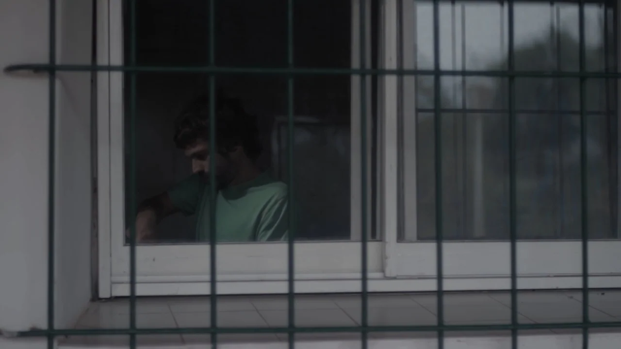 Still Life (2013) movie still: medium — A man stands inside a room visible through a window frame with metal bars. The man looks down while wearing…