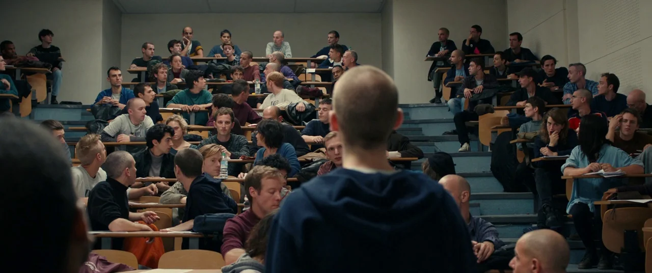 BPM (Beats Per Minute) (2017) movie still: medium — A tiered university lecture hall filled with many students seated at wooden desks. A student…