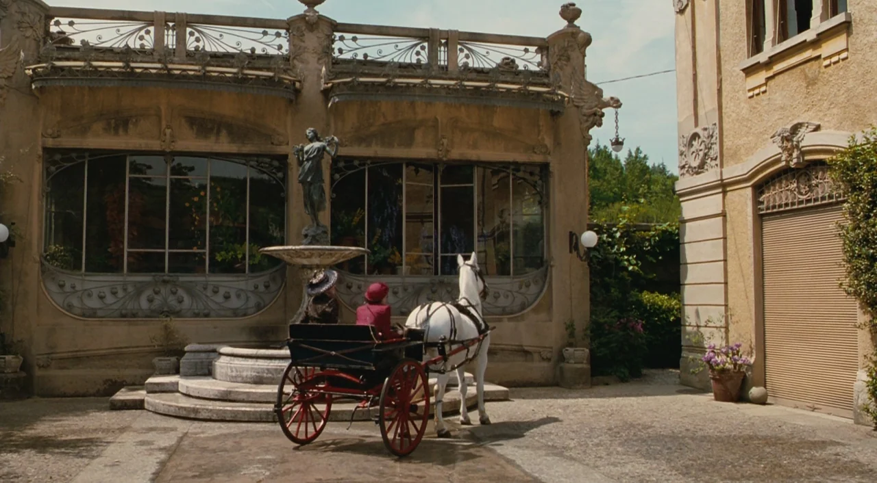 Life is Beautiful (1997) movie still: wide — A stone courtyard features an ornate building with glass windows and a central fountain. Two people sit…