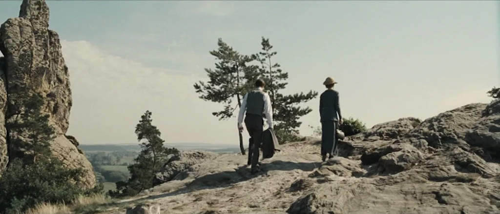 Frantz (2016) movie still: wide — A rocky mountain plateau overlooking a valley with scattered pine trees. A man and a woman walk across the uneven…