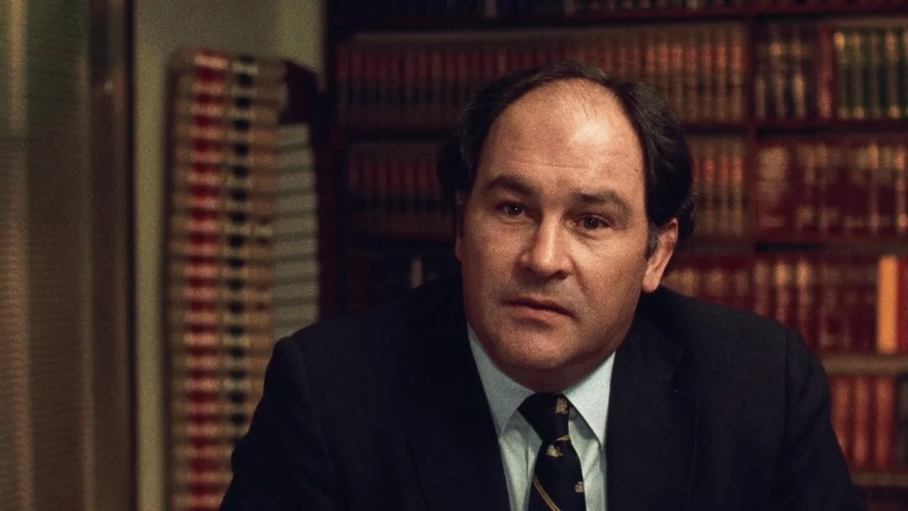The Thin Blue Line (1988) movie still: medium — A man sits in front of a wall lined with many leather-bound books. He faces the camera while wearing…