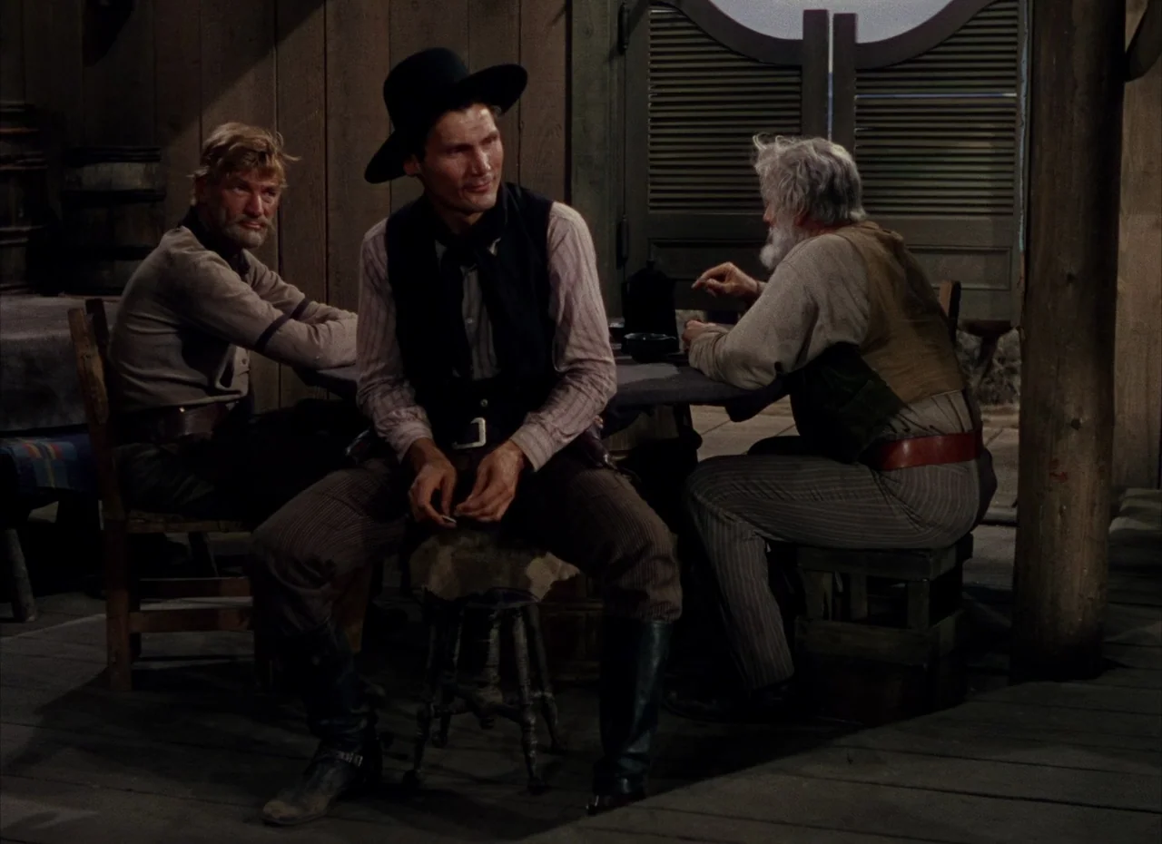 Shane (1953) movie still: medium — A dimly lit saloon features dark wood walls and saloon-style swinging doors at back. Three men sit around a small…