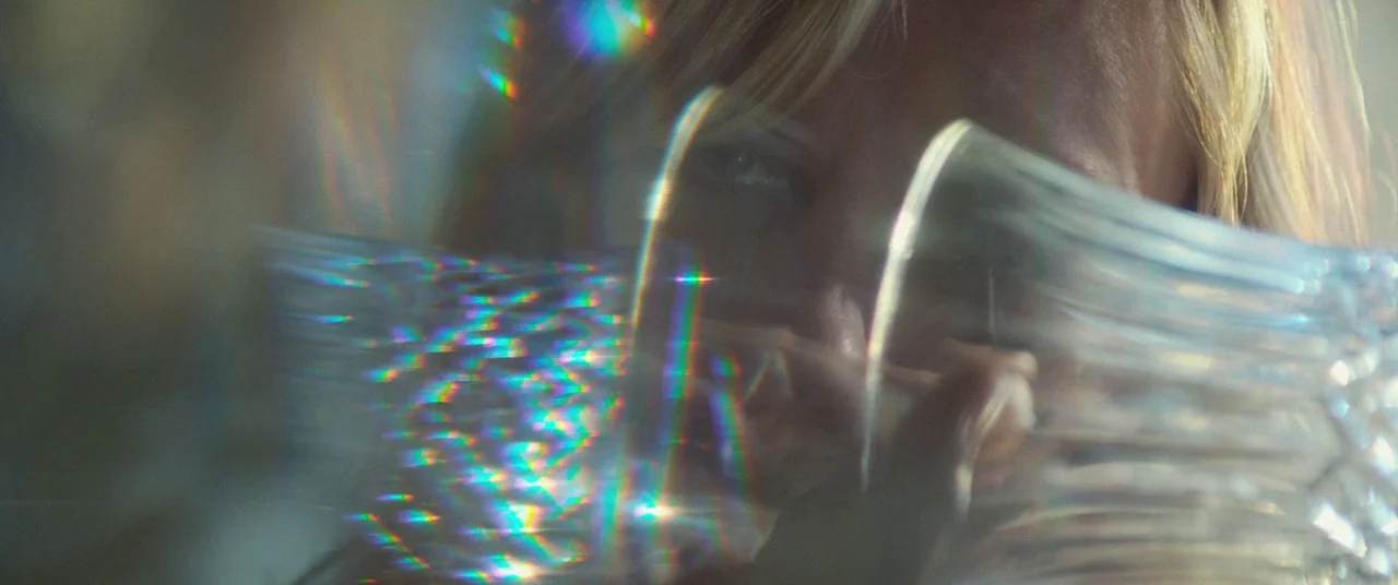 Woodshock (2017) movie still: closeup — A woman's face is partially obscured by a textured clear glass object. Bright rainbow light refractions from…
