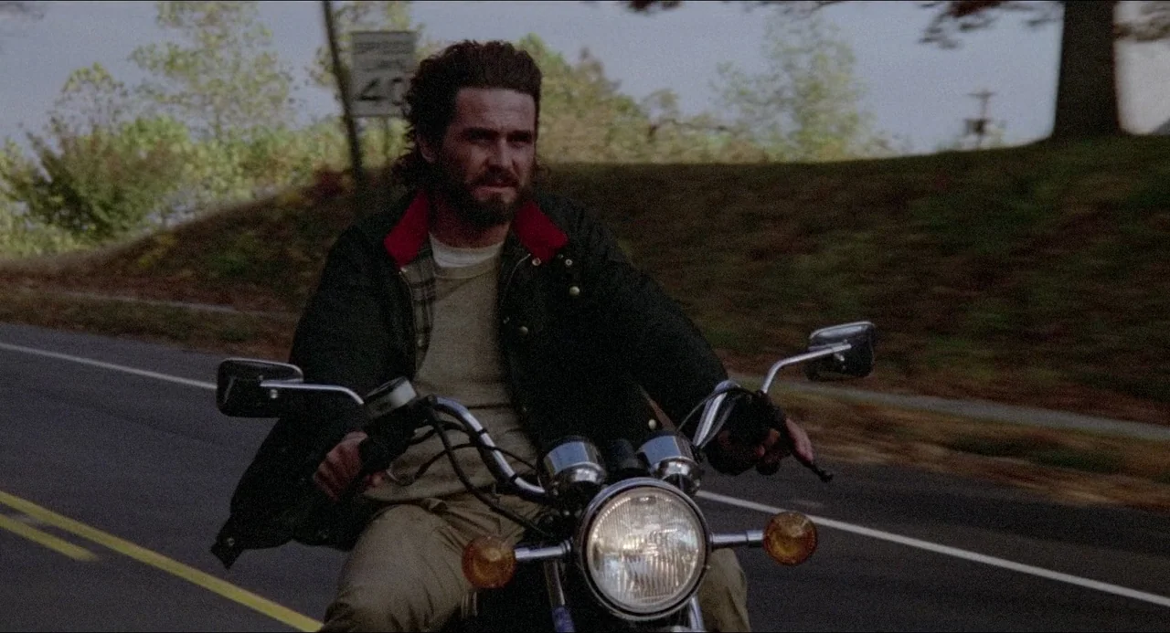 The Amityville Horror (1979) movie still: medium — A man rides a motorcycle along a paved road flanked by grassy slopes. The road has a painted…