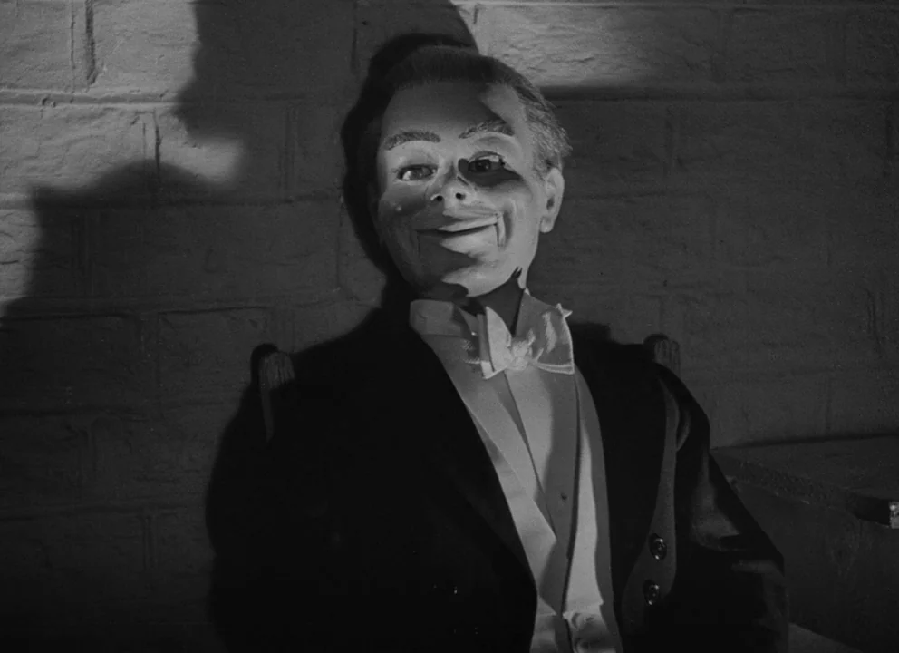 Dead of Night (1945) movie still: medium — A ventriloquist dummy sits against a textured brick wall in a dimly lit space. The puppet wears a tuxedo…