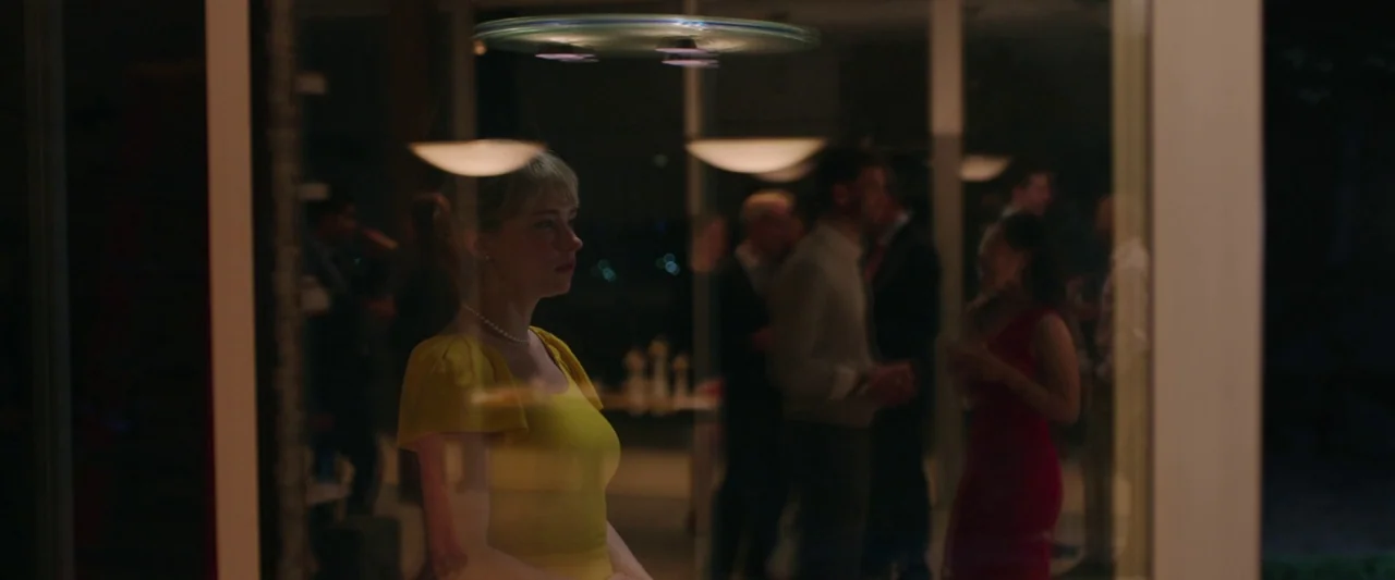 Swallow (2020) movie still: medium — A woman stands outside a modern glass-walled structure containing a social gathering at night. The woman in the…