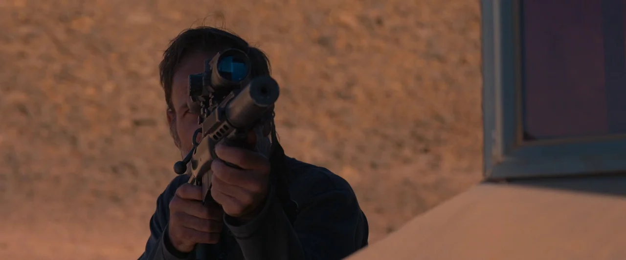 Settlers (2021) movie still: medium — A rugged desert landscape serves as the backdrop for an armed individual. A man with a thick beard holds a…