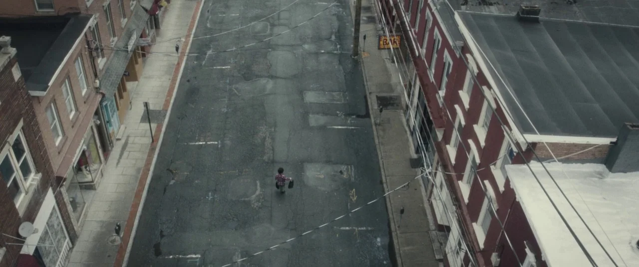 I Think We’re Alone Now (2018) movie still: wide — An empty urban street lined with brick commercial buildings is viewed from above. A small child…