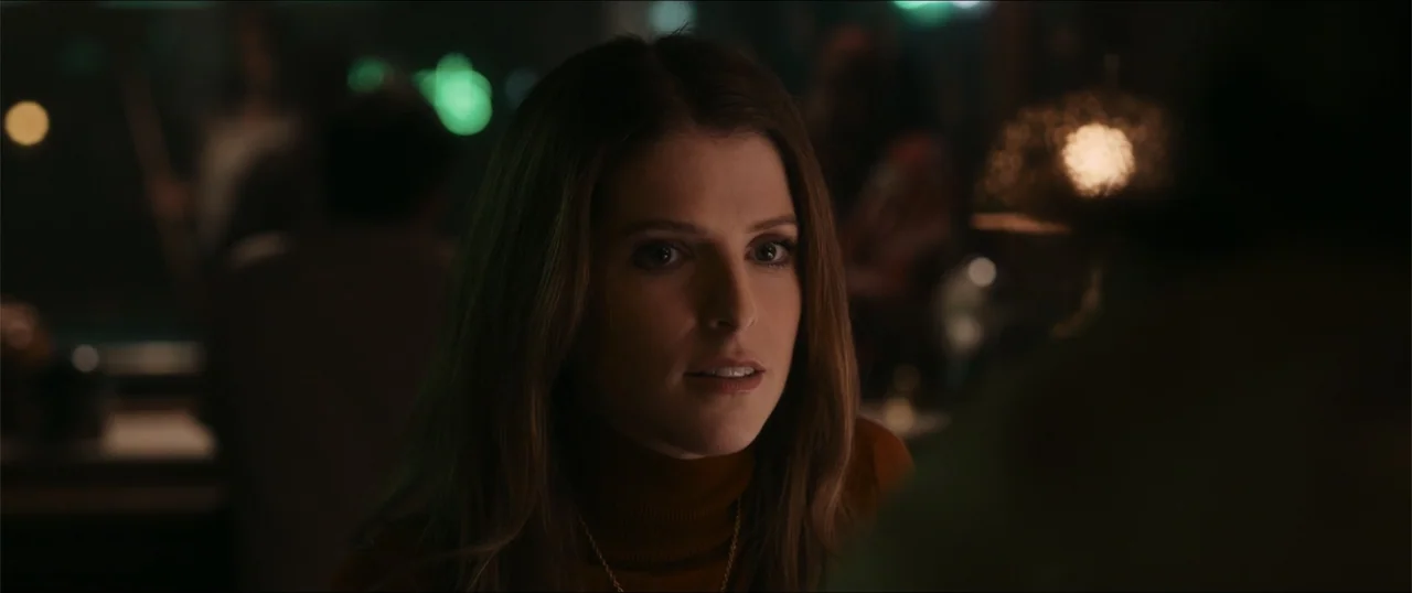 Woman of the Hour (2024) movie still: closeup — A dimly lit bar interior featuring soft out-of-focus background lights. A woman in a dark turtleneck…