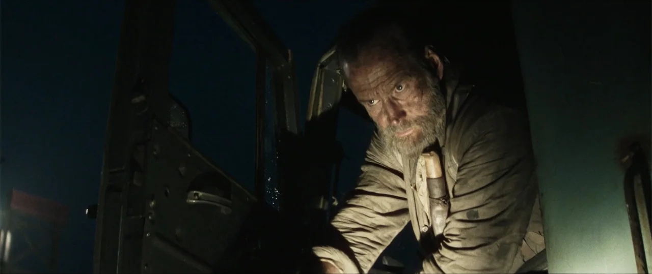 Sisu: Road to Revenge (2025) movie still: medium — A darkened vehicle cab interior features a man leaning toward an open door. The man peers into…