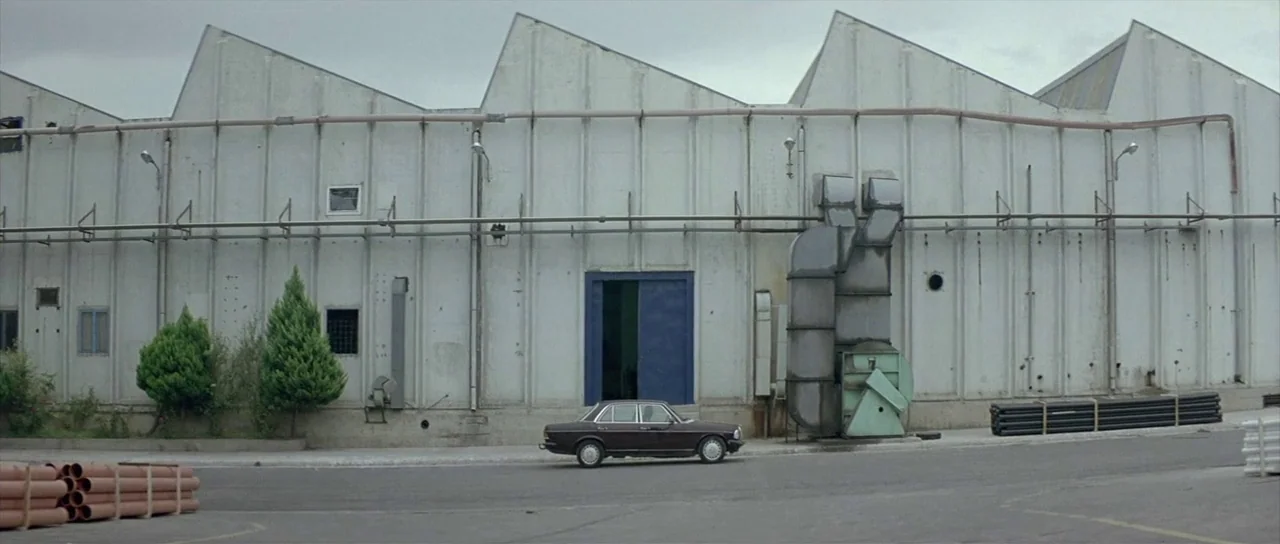 Dogtooth movie still: wide — A long white industrial building features a sawtooth roof and blue sliding doors. A dark-colored vintage sedan sits…