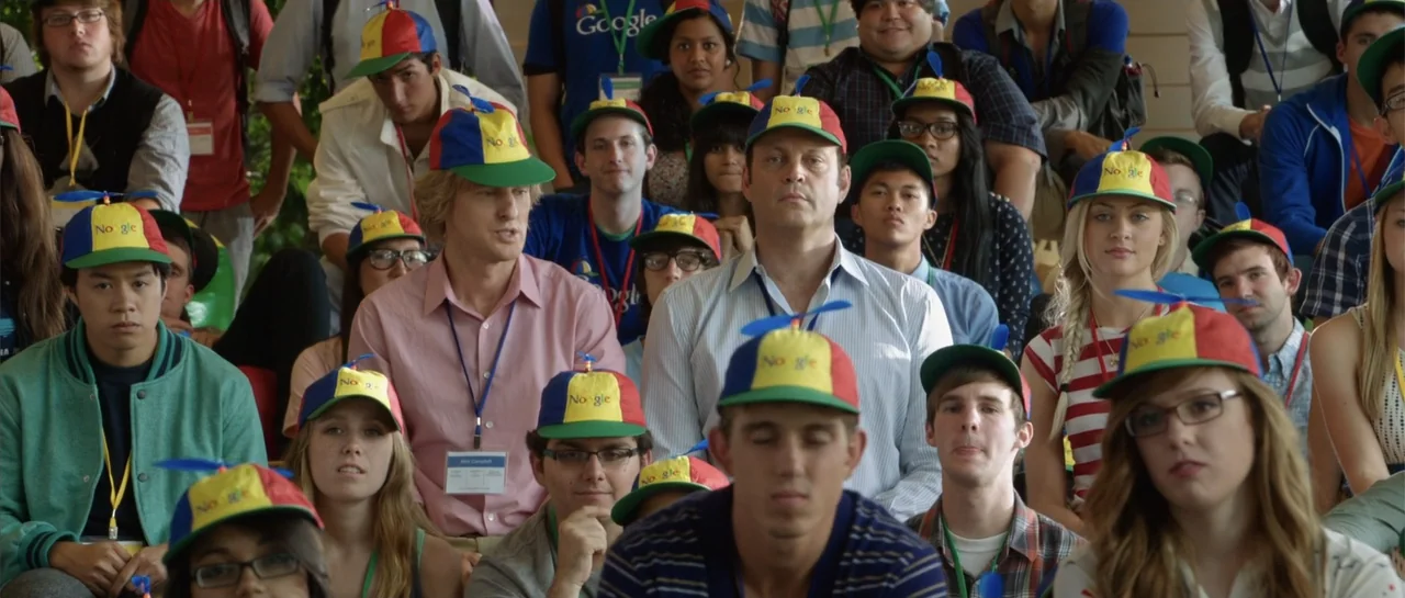 The Internship (2013) movie still: medium — A crowded room contains many people sitting in rows while wearing colorful propeller hats. The subjects…