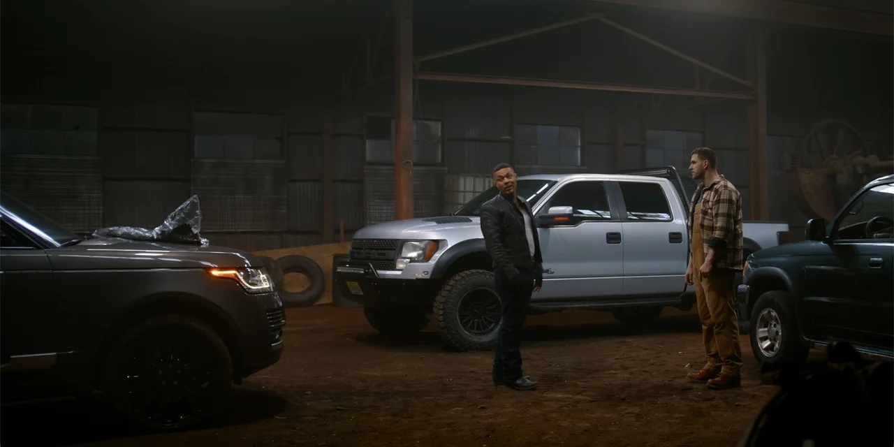 The Pickup (2025) movie still: fullbody — A dimly lit industrial space features several vehicles parked on a dirt floor. Two men stand facing each…