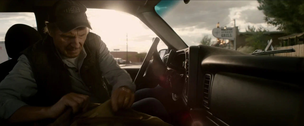 The Marksman (2021) movie still: medium — A man sits in the driver seat of a vehicle during the late afternoon. He reaches into a bag placed on his…