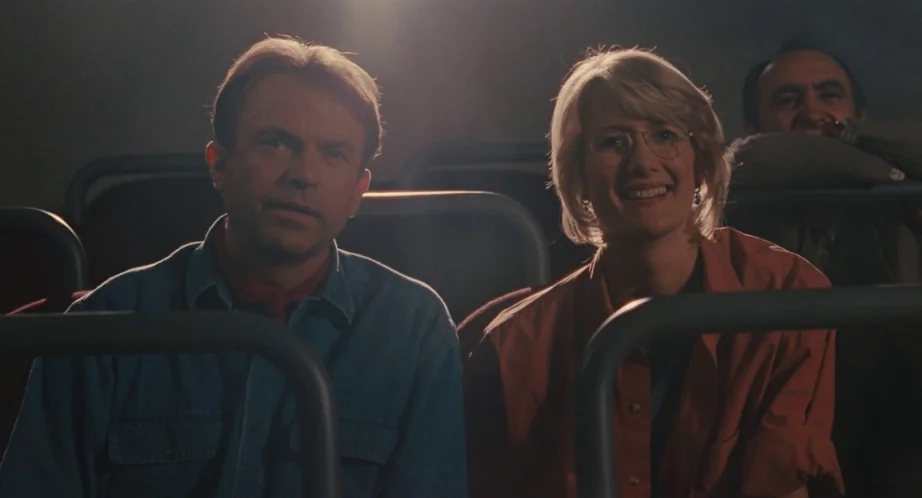 Jurassic Park (1993) movie still: medium — Two individuals sit in dark, padded cinema seats inside a dimly lit auditorium. A man and a woman look…