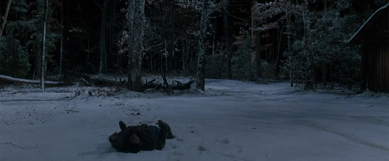 Fallen (2004) movie still: wide — A dark, snow-covered clearing sits surrounded by a dense, leafless forest at night. A person lies flat on their…