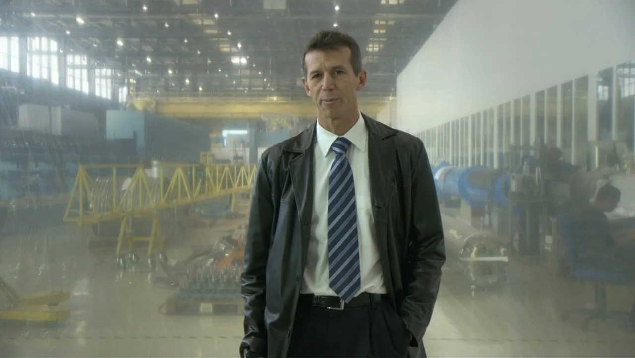 CERN (2013) movie still: medium — A spacious industrial manufacturing hall contains machinery and equipment behind a plastic curtain. A middle-aged…