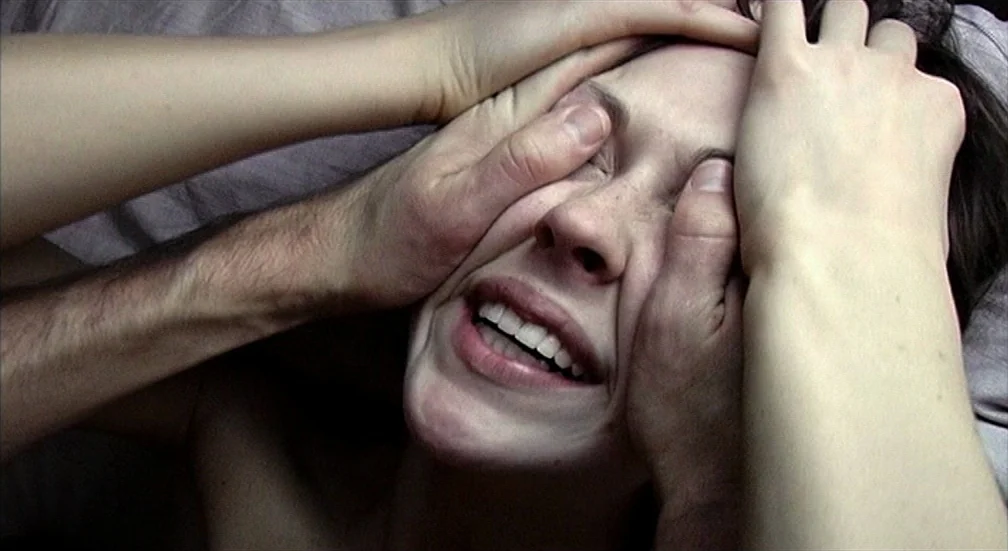 9 Songs (2004) movie still: closeup — A person lies back on a grey fabric surface with their eyes covered. Someone's hands firmly grip the sides of…