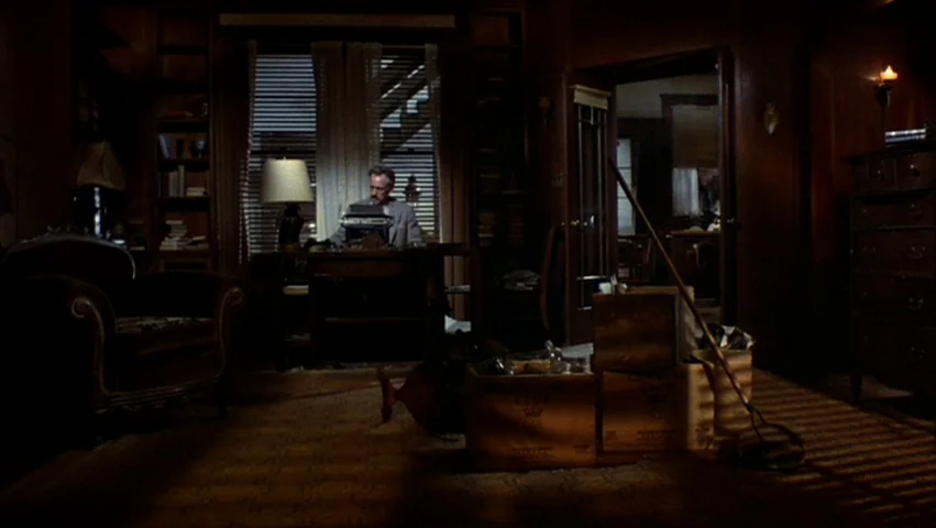 Hammett (1982) movie still: wide — A dimly lit home workspace features wooden furniture, book-filled shelves, and stacked cardboard boxes. A man…