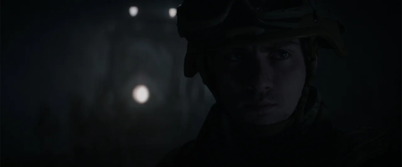 Godzilla (2014) movie still: closeup — A dark night scene features a soldier in tactical gear standing in the shadows. A single blurred light source…