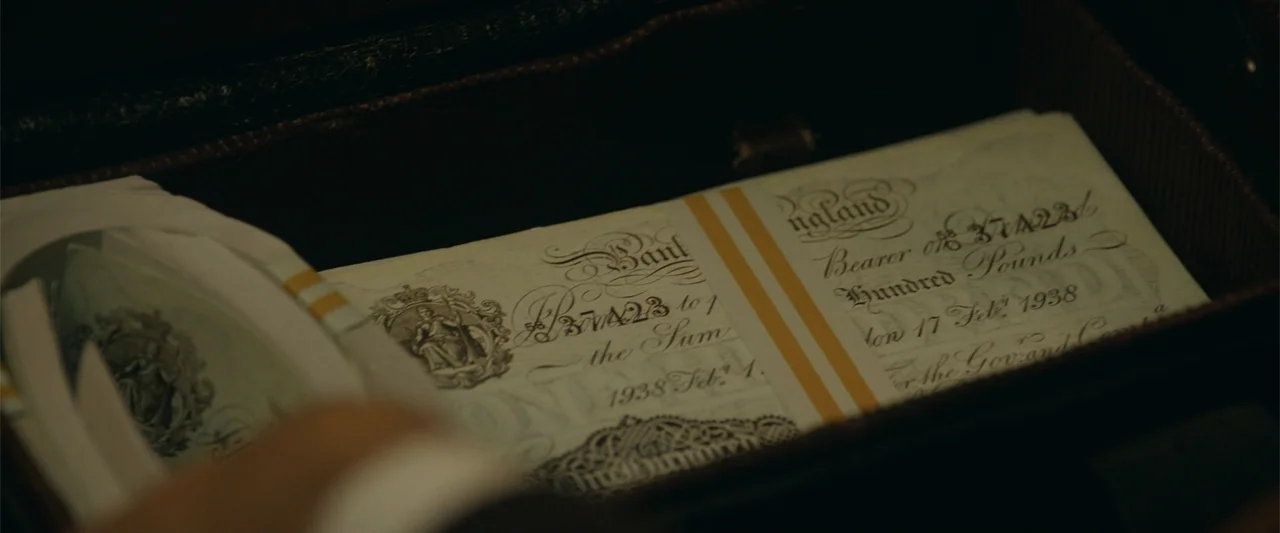 Sardar Udham (2021) movie still: closeup — An open dark leather briefcase holds a stack of vintage British hundred-pound banknotes. The paper money…