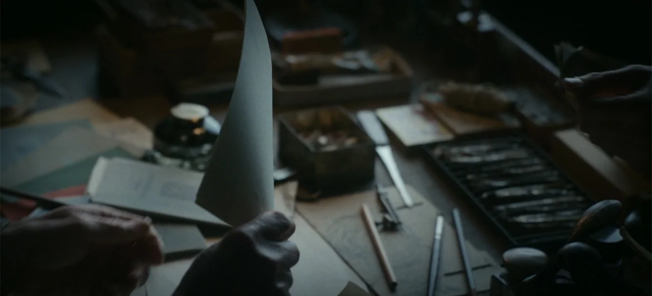 One Life (2023) movie still: closeup — A cluttered wooden drafting desk holds various drawing tools, an inkwell, and paper. Hands hold a single…