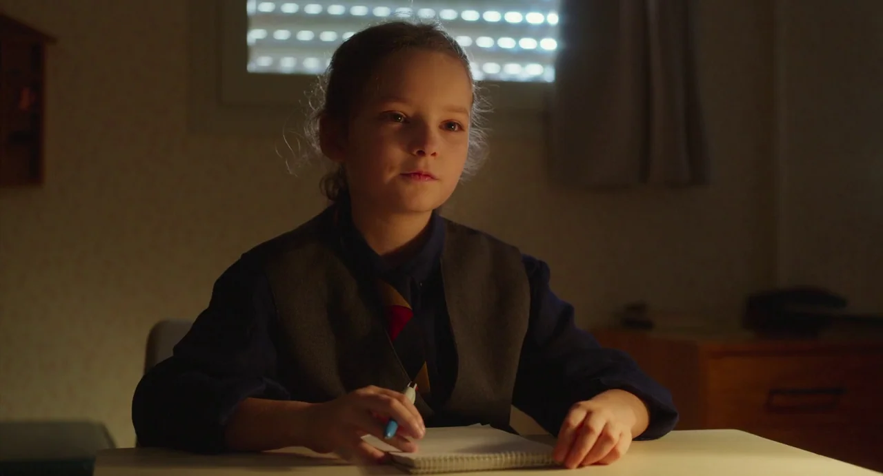 Petite Maman (2021) movie still: medium — A dimly lit room features a student sitting at a table near a window. A young girl in school uniform sits…