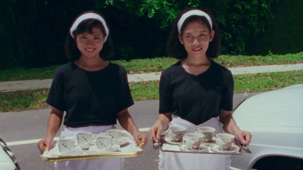 Shirkers (2018) movie still: medium — Two women wearing white headbands and matching uniforms stand on a suburban street. Each woman carries a…