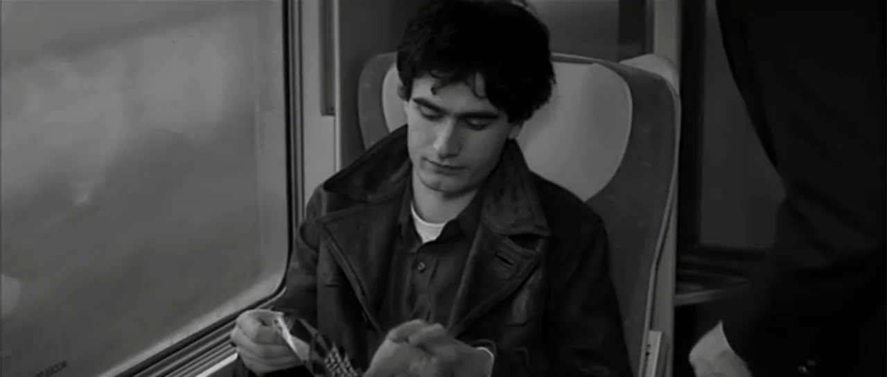 13 Tzameti (2006) movie still: medium — A passenger compartment on a vintage train with large rectangular windows and patterned seating. A young man…