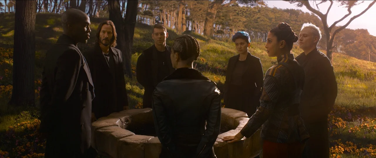 The Matrix Resurrections (2021) movie still: fullbody — Seven people stand in a ring around an old stone well in a forest. The group is surrounded…