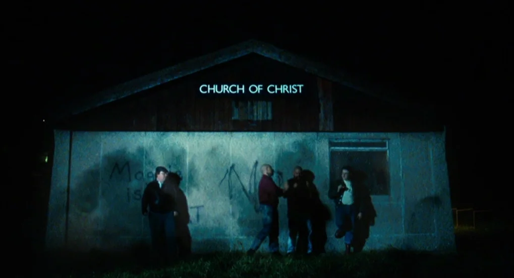 This Is England (2006) movie still: wide — A small, dilapidated building with a brightly illuminated neon sign above the entrance. Four men stand…