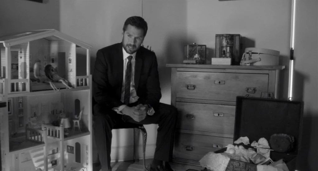 Much Ado About Nothing (2012) movie still: medium — A man in a suit sits in a room next to a large dollhouse. A chest of drawers and an open…