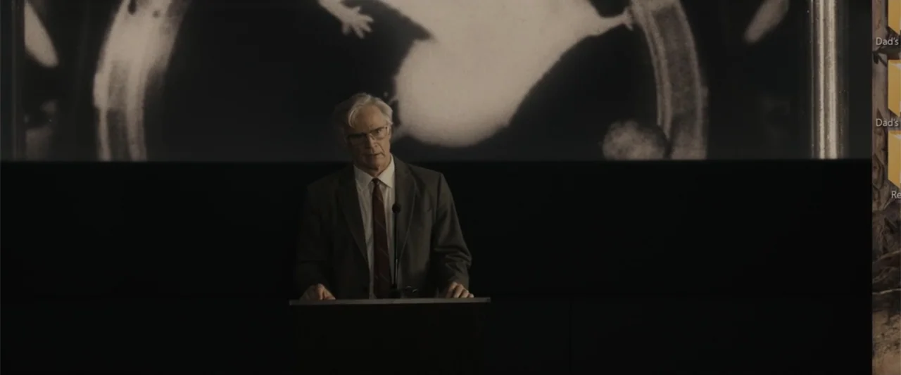 Afraid (2024) movie still: medium — A dark lecture hall features a large projection screen displaying a black and white image. An older man in a…