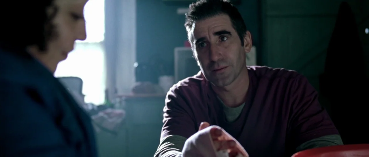 Isolation (2005) movie still: medium — A dimly lit room with blue walls and a window letting in soft daylight. A man with a bandaged hand sits at a…