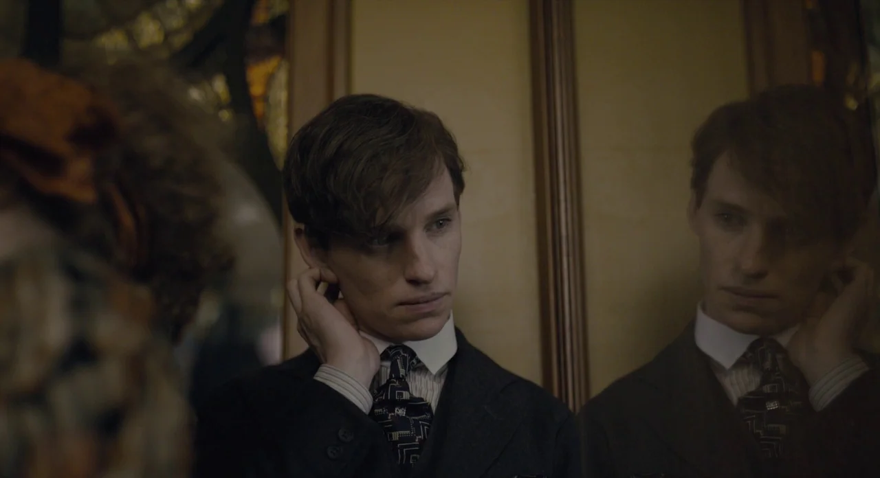 The Danish Girl (2015) movie still: medium — An interior space with wood paneling features a reflective surface showing a young man. A man in a suit…