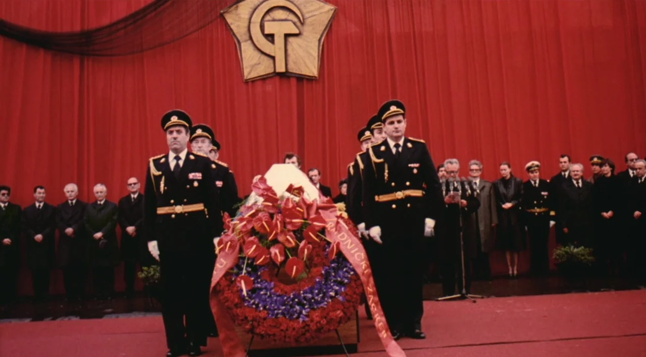 Underground (1995) movie still: fullbody — A formal stage features a prominent gold communist-style symbol against a massive red curtain. Two men in…