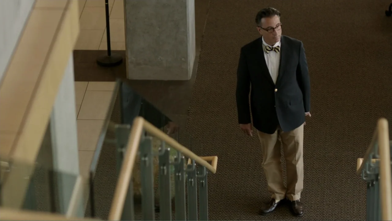 At Middleton (2013) movie still: fullbody — A man stands on a carpeted landing near an office staircase with glass railings. The man looks toward…