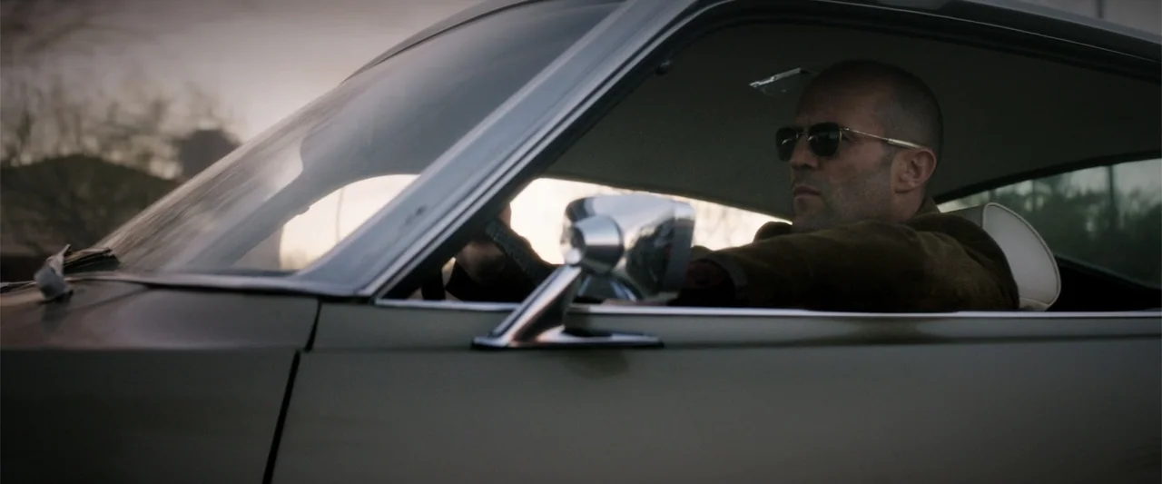 Wild Card (2015) movie still: medium — A man drives a vintage car through an outdoor setting during golden hour. The driver wears sunglasses and a…