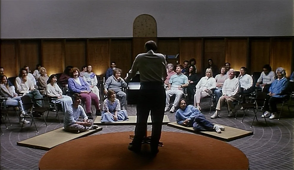 Safe movie still: wide — A spacious room with wood paneling and a circular rug serves as a meeting area. A group of diverse adults sit in chairs and…