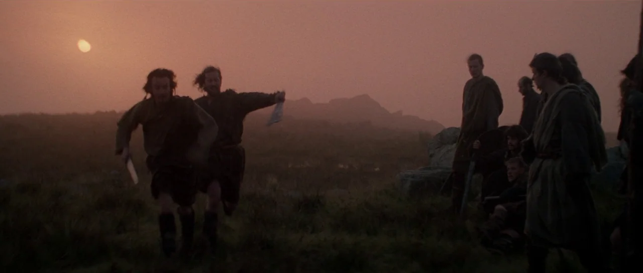 Braveheart (1995) movie still: wide — An open field at dusk with rolling hills silhouetted against a setting sun. Several men stand in a group as…