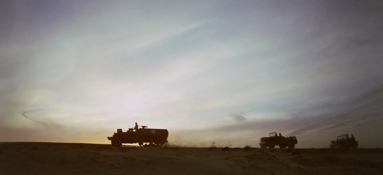 Patton (1970) movie still: wide — An open desert landscape stretches toward a pale, cloudy horizon at sunset. Three military vehicles drive in a…