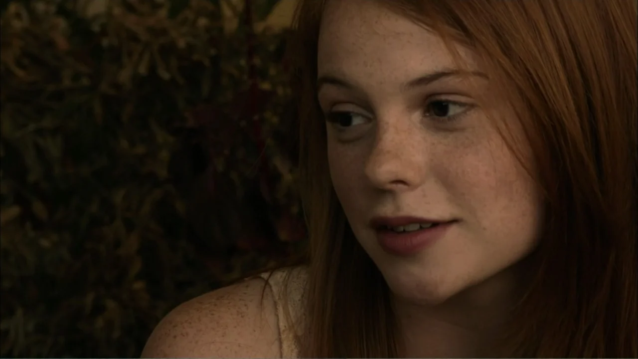 Dark Skies (2013) movie still: closeup — A dimly lit interior room with dark, out-of-focus foliage visible in the background. A young woman with red…