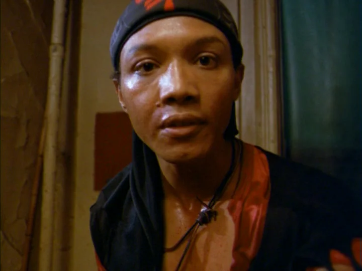 Tenement (1985) movie still: closeup — A cramped and dim corridor with off-white walls and visible vertical piping. A man wearing a black bandana…