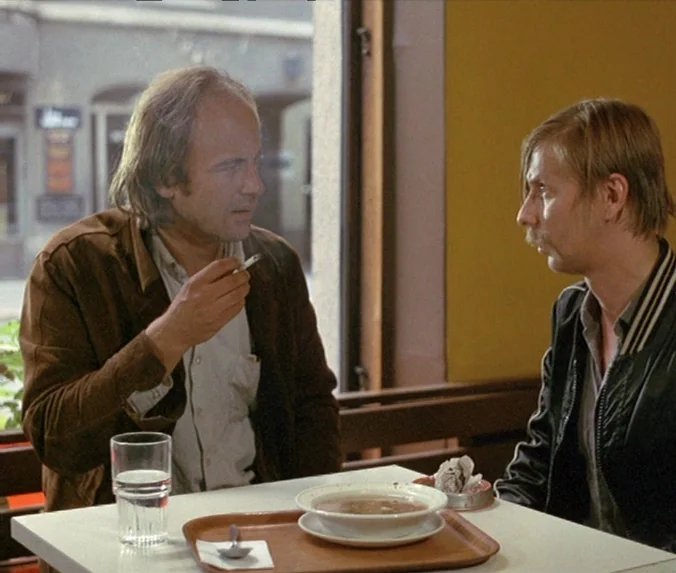 Crime & Punishment (1983) movie still: medium — A modest dining area features a simple white table set against a yellow wall. Two men sit across…