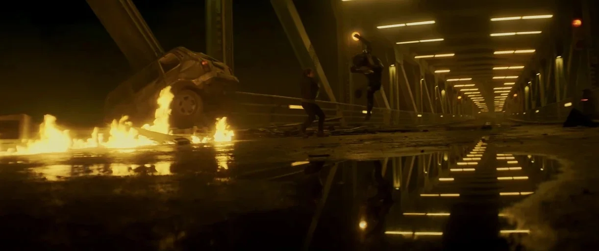 Black Widow (2021) movie still: wide — A metallic bridge at night features a burning car wreck and rows of overhead lights. One person kicks at…