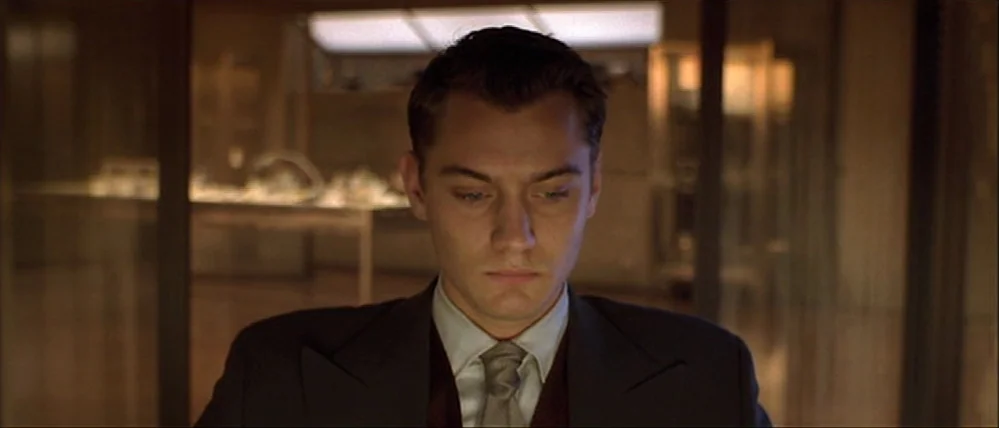 Gattaca (1997) movie still: closeup — A dark, modern office space contains glass partitions and minimalist furniture. A man in a suit looks downward…