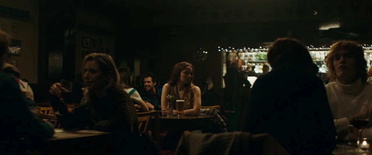 Saint Maud (2019) movie still: medium — A dimly lit room with multiple small wooden tables and various patrons gathered inside. Several people sit…